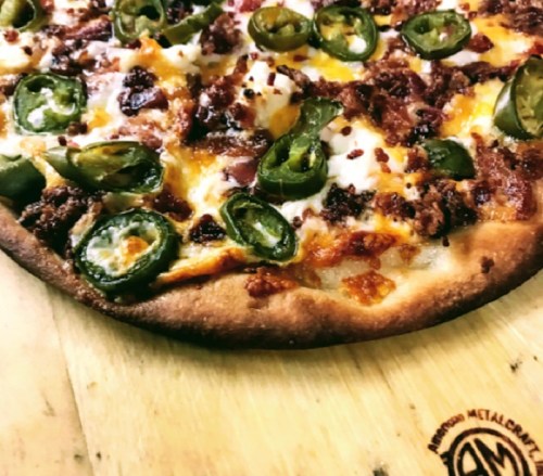 Close-up of a pizza topped with jalapeños, bacon, and melted cheese on a golden crust, served on a wooden board.