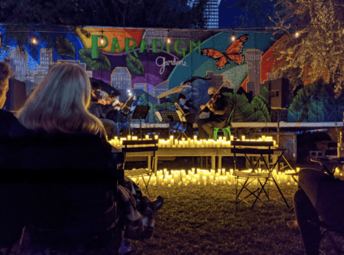 A nighttime outdoor concert with musicians performing in front of a colorful mural, surrounded by glowing candles.