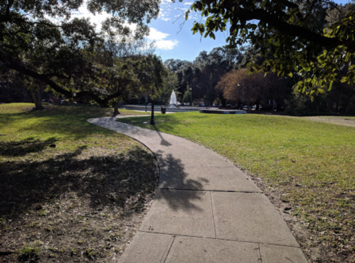 A winding pathway through a park, with trees and a fountain visible in the background under a clear blue sky.