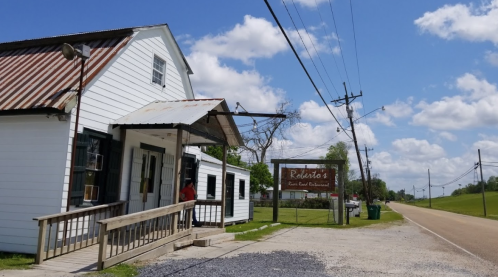 A rustic white building with a metal roof, featuring a sign for "Roberto's Restaurant" and a gravel path beside a road.