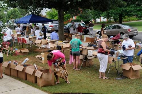 A community yard sale with people browsing and sorting through items in cardboard boxes on a grassy area.