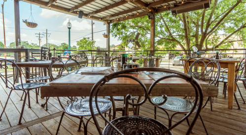 Outdoor dining area with wooden tables and metal chairs, surrounded by greenery and a sunny sky.