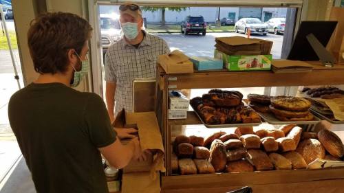 A masked customer orders baked goods from a vendor behind a display of various breads and pastries.