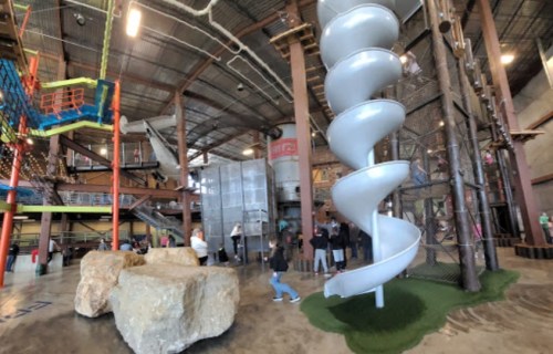 Indoor play area featuring a spiral slide, climbing structures, and large rocks, with children and adults interacting.