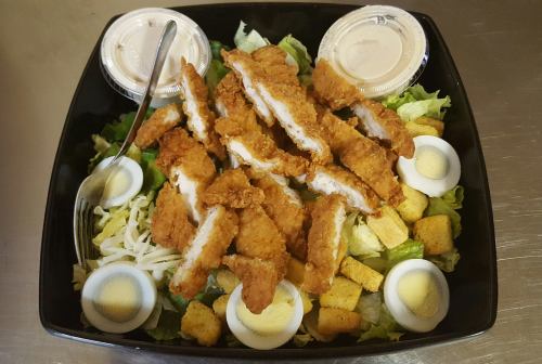 A salad topped with crispy chicken strips, sliced hard-boiled eggs, croutons, and two dressing cups.