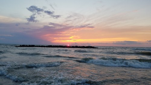 Sunset over the ocean, with waves gently lapping at the shore and a rocky jetty silhouetted against the colorful sky.