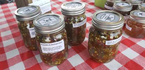 Jars of candied jalapeños on a red and white checkered tablecloth at a market. Labels detail ingredients and usage.