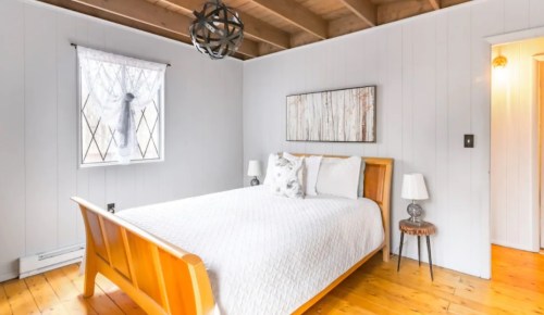 Cozy bedroom with a wooden bed, white bedding, and natural light from a window, featuring minimalist decor.