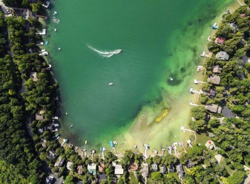 Aerial view of a serene lake surrounded by lush greenery and homes, with boats on the water.