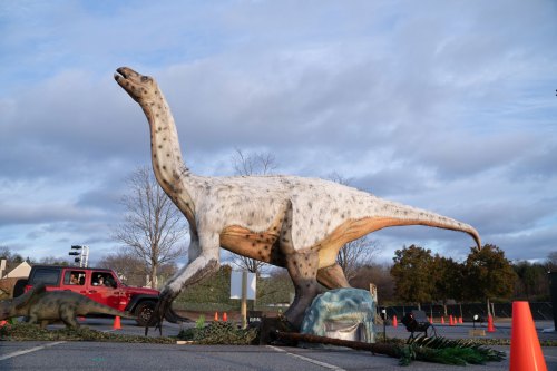 A large dinosaur statue stands on a road, with a red jeep nearby and trees in the background under a cloudy sky.