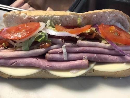 A close-up of a sub sandwich filled with layers of deli meats, cheese, lettuce, tomatoes, and onions on a hoagie roll.