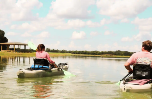 Two people kayaking on a calm lake, surrounded by greenery and a blue sky with fluffy clouds.