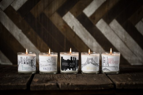 Five lit candles with city-themed designs on a rustic wooden surface, set against a patterned wooden background.