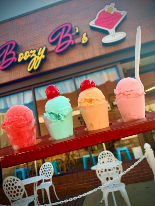 Four colorful ice cream scoops in cups, topped with cherries, in front of a shop called Boozy B's.