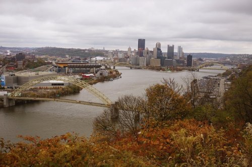 A panoramic view of Pittsburgh's skyline with rivers and bridges, surrounded by autumn foliage under a cloudy sky.