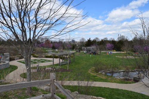 A scenic park with pathways, trees, and colorful play areas under a partly cloudy sky.