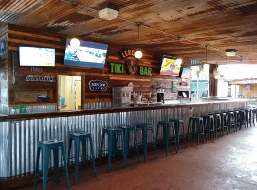 Interior of a tiki bar with a metal bar top, stools, and multiple TVs displaying sports. Rustic wooden decor.