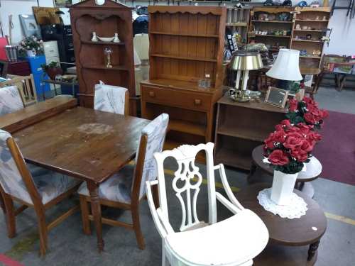 A variety of vintage furniture including tables, chairs, and shelves, with decorative flowers in a shop setting.