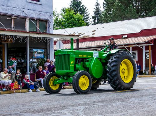 A green and yellow vintage tractor drives past a crowd during a parade, with people seated along the street.