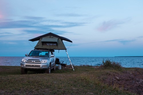 A silver SUV with a rooftop tent parked by the shore, overlooking a calm lake at dusk.