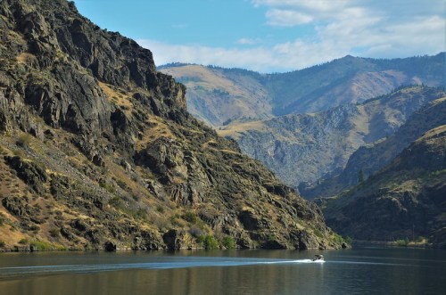 A serene river flows between rugged mountains under a blue sky with scattered clouds. A boat glides across the water.