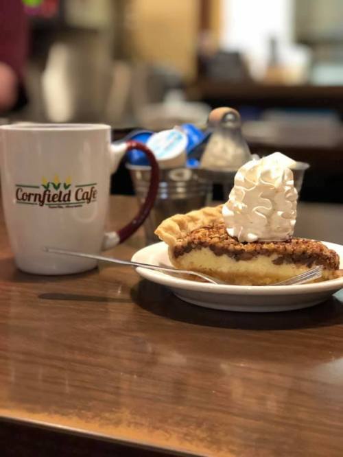 A slice of pie topped with whipped cream on a plate, next to a coffee mug from Cornfield Cafe.