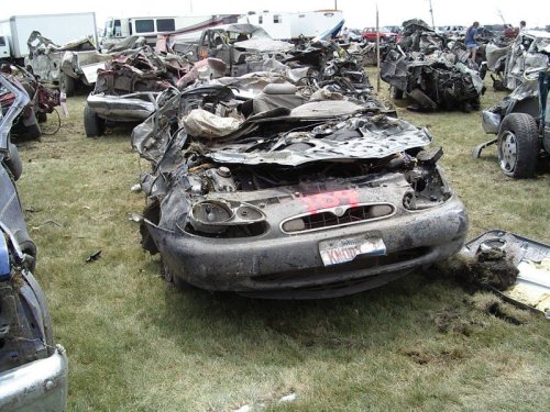 A damaged, rusted car in a junkyard, surrounded by other wrecked vehicles and overgrown grass.