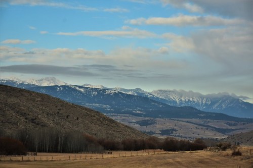 A scenic view of snow-capped mountains under a blue sky, with rolling hills and a grassy field in the foreground.