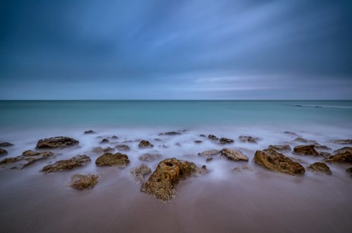 A serene seascape featuring smooth rocks in shallow water under a moody, overcast sky.