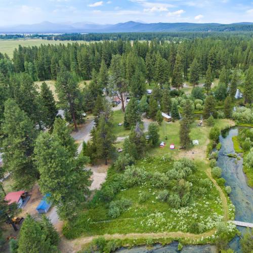 Aerial view of a lush campsite surrounded by trees, with tents and a winding stream in a scenic landscape.