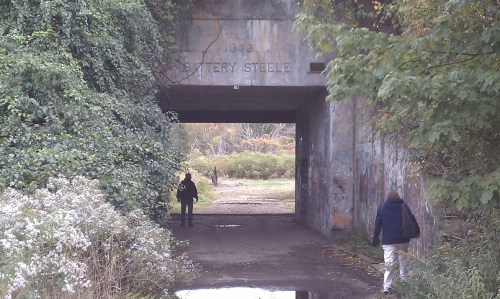 Two people walk through an overgrown tunnel marked "Battery Steele," surrounded by greenery and graffiti.