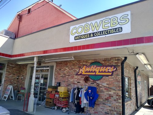 Exterior of a store named "Cobwebs Antiques & Collectibles," featuring a brick facade and vintage items for sale outside.
