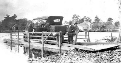 A vintage photo of men by a wooden dock with an early car, surrounded by water and trees.