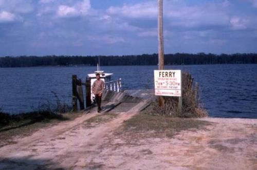 A man walks along a dirt path by a river, near a sign for a ferry service and a small boat docked nearby.