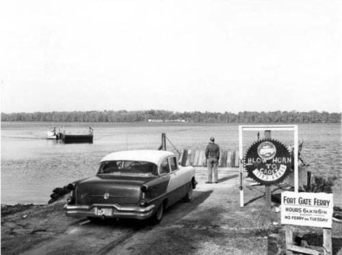 A vintage car parked near a ferry dock, with a sign indicating ferry hours and a boat approaching across the water.