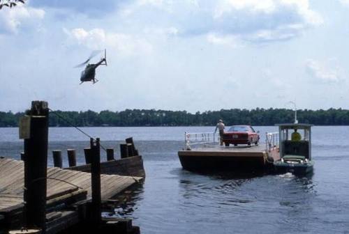 A helicopter flies over a river as a car drives onto a ferry, with a dock visible in the foreground.