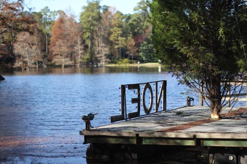 A wooden dock by a lake with a sign reading "NOEL" and trees in the background reflecting autumn colors.