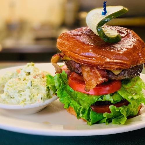 A juicy burger with bacon, lettuce, tomato, and a pickle on a toasted bun, served with a side of coleslaw.