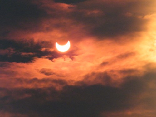 A partially eclipsed sun peeks through dark, swirling clouds, casting a warm orange glow in the sky.