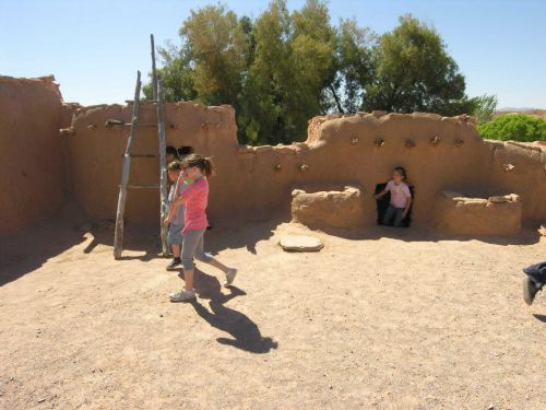 Children exploring a historic adobe structure with a ladder and a small entrance, surrounded by trees and clear blue sky.