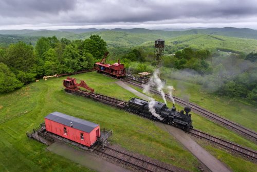 A vintage steam train puffs smoke near a red train car and machinery, surrounded by lush green hills under a cloudy sky.