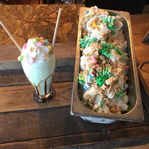 A milkshake topped with whipped cream and colorful sprinkles next to a tray of green and pastel-colored dessert.