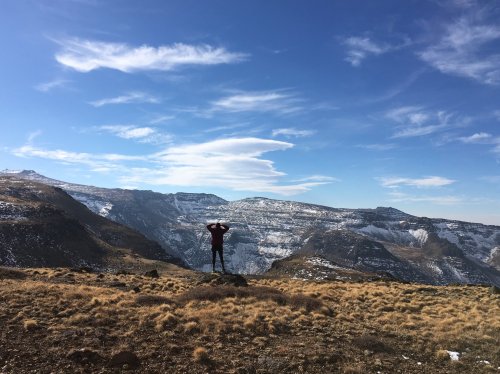A person stands on a rocky outcrop, arms raised, overlooking a vast mountainous landscape under a blue sky.