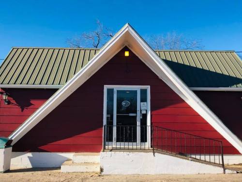 A red A-frame building with a green metal roof and a front door, set against a clear blue sky.