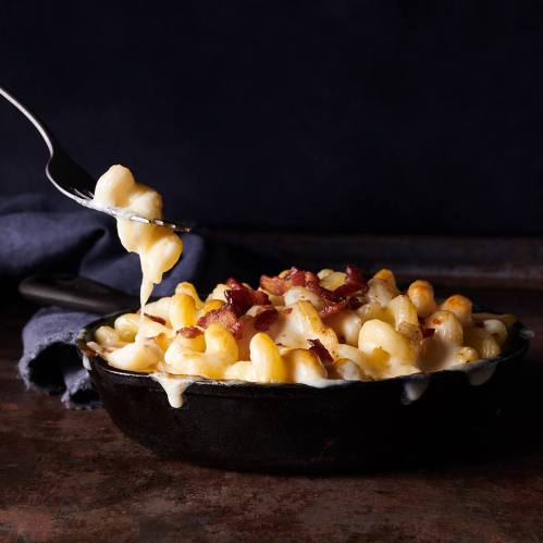 A fork lifts creamy macaroni and cheese topped with crispy bacon from a cast iron skillet.