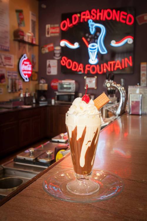 A classic soda fountain dessert with whipped cream, a cherry, and a wafer, set against a neon "Old Fashioned Soda Fountain" sign.