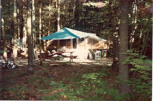 A tent campsite surrounded by trees, featuring a blue canopy, picnic table, and camping gear.