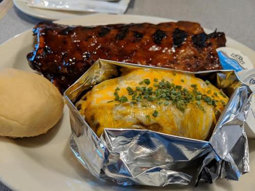 A plate featuring grilled ribs, a cheesy baked potato wrapped in foil, and a soft dinner roll.
