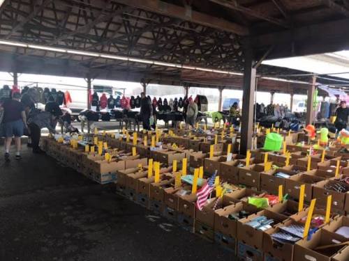 A bustling market with rows of boxes filled with items, under a covered area, and shoppers browsing around.
