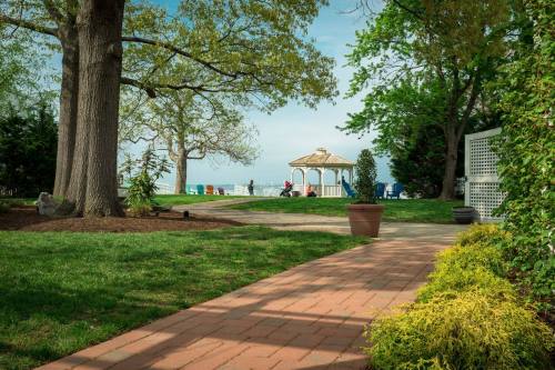 A serene garden path leads to a gazebo by the water, surrounded by trees and colorful chairs.